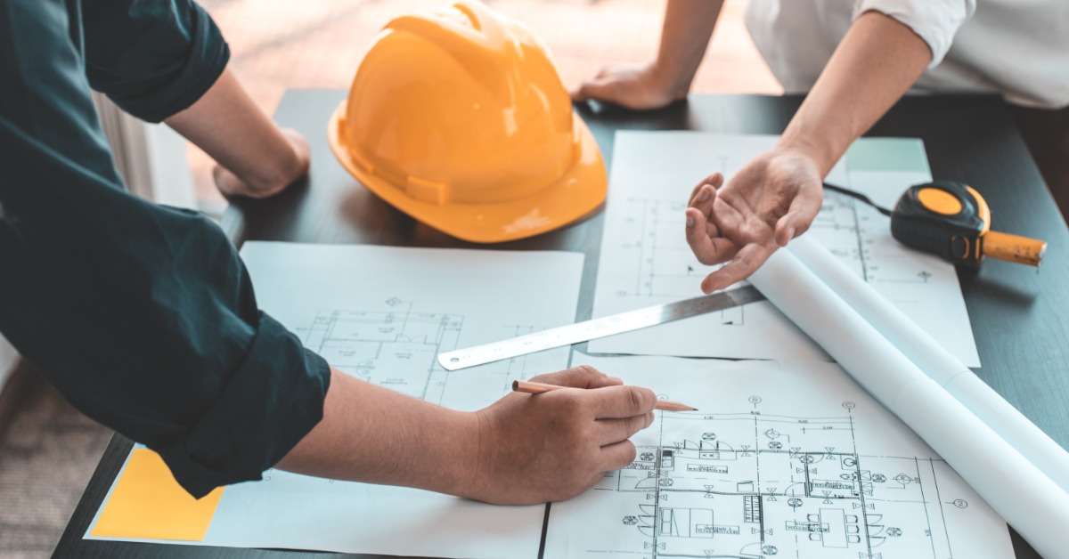 lesterbuildingsystems-470710-people-building-plans-imageb.jpg People review building plans at a table with a hard hat, measuring tape, and rolled construction drawings.