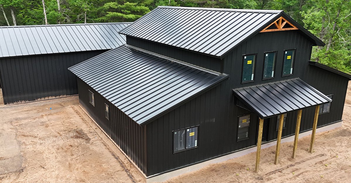 Lester-Building-Systems-LLC-439188-ImageA.jpg A black metal building with a gray roof, covered entry, concrete drive, green windows, and trees behind it.