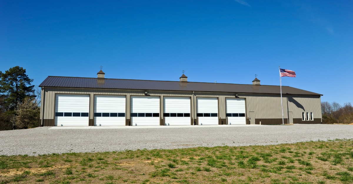 lesterbuildingsystemsllc-470705-rural-fire-station-imageb.jpg A large commercial building features six overhead garage doors, a metal exterior, cupolas, and an American flag.