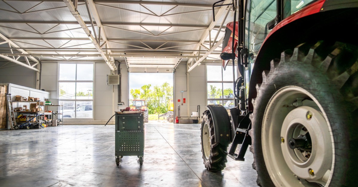 lesterbuildingsystems-470700-tractor-shop-tools-imagea.jpg A tractor sits inside a spacious farm shop with an open work area, tool cart, shelving, and overhead lighting.