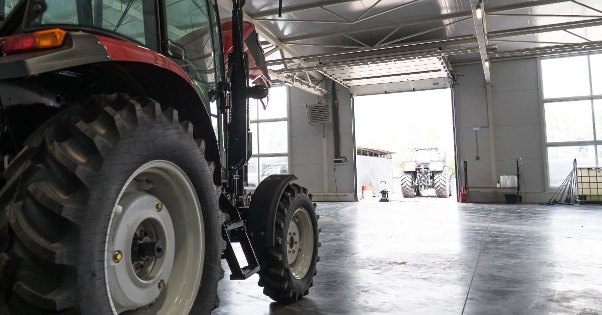lesterbuildingsystems-470714-tractor-storage-building-imagea.jpg A red tractor sits on a concrete floor inside a farm workshop with an open overhead door and another tractor outside.