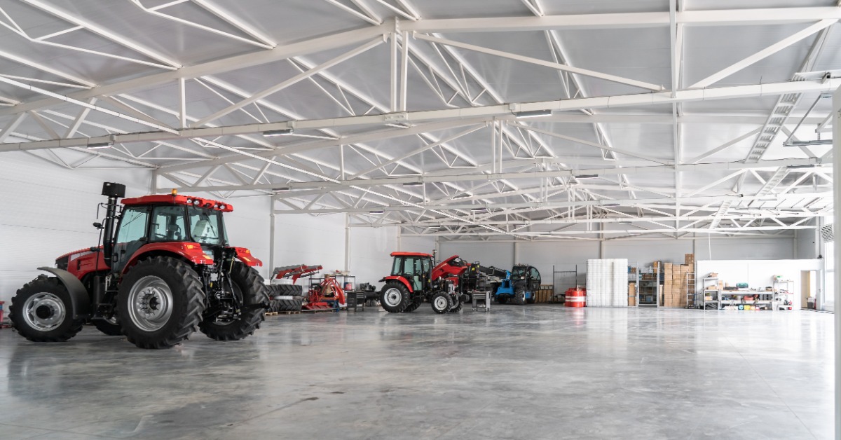 Several red tractors and farm machines sit inside a large building with white roof trusses and an open concrete floor.