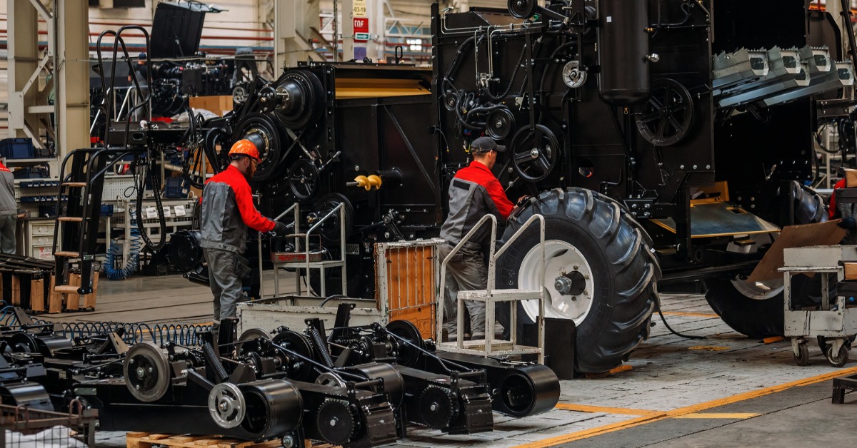 lesterbuildingsystemsllc-470707-workers-repair-equipment-imagea.jpg Two workers assemble a large black agricultural machine with tires and mechanical components in a factory.