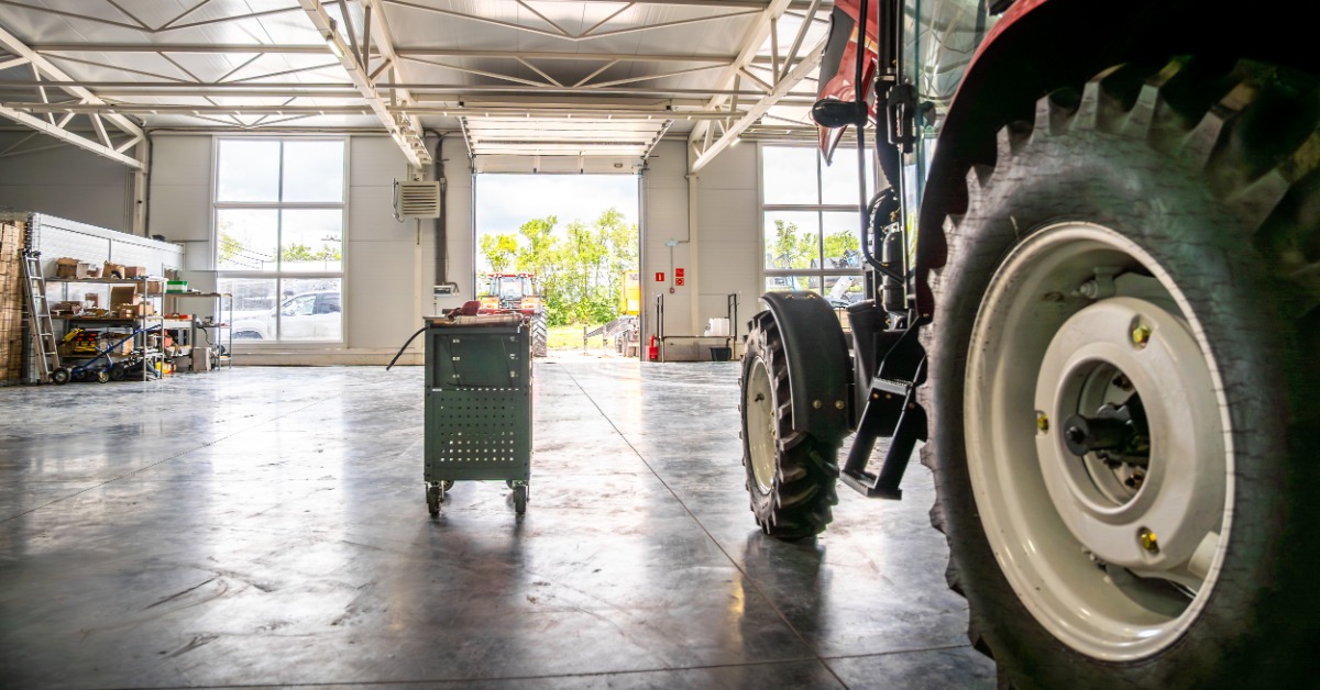 lesterbuildingsystemsllc-470707-interior-tractor-workshop-imageb.jpg A red tractor is inside a farm workshop with a rolling tool cart, an open overhead door, shelving, and a concrete floor.