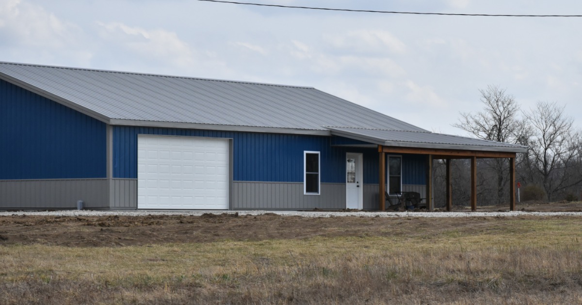 A blue and gray barndominium features an attached garage, covered porch, and residential entrance on a rural property.