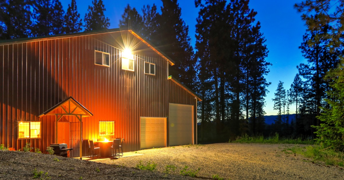 A dark metal barndominium with garage doors and a lit residential entrance stands among tall trees at dusk.