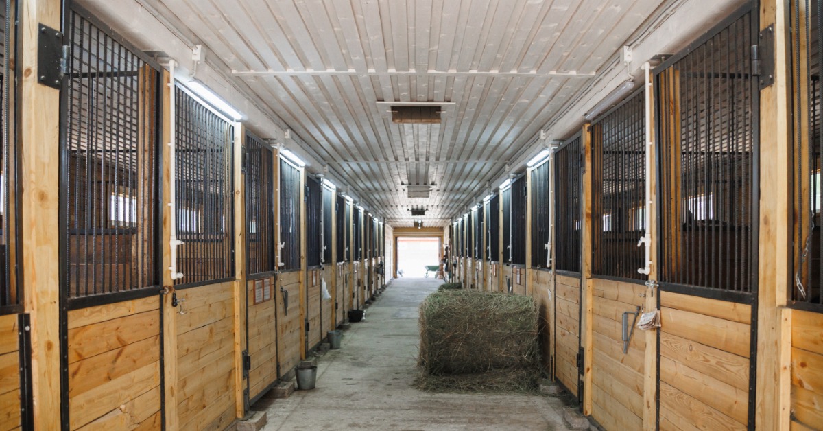 A long barn aisle with wooden horse stalls on both sides, featuring metal gates, overhead lighting, and a bale of hay.